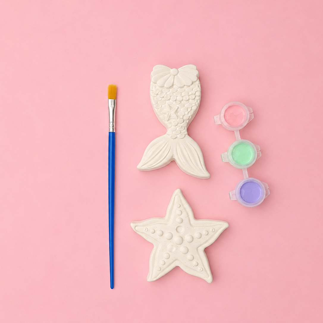Decorative items including a star-shaped object, a mermaid tail, paintbrush, and small bottles of paint on a pink background.